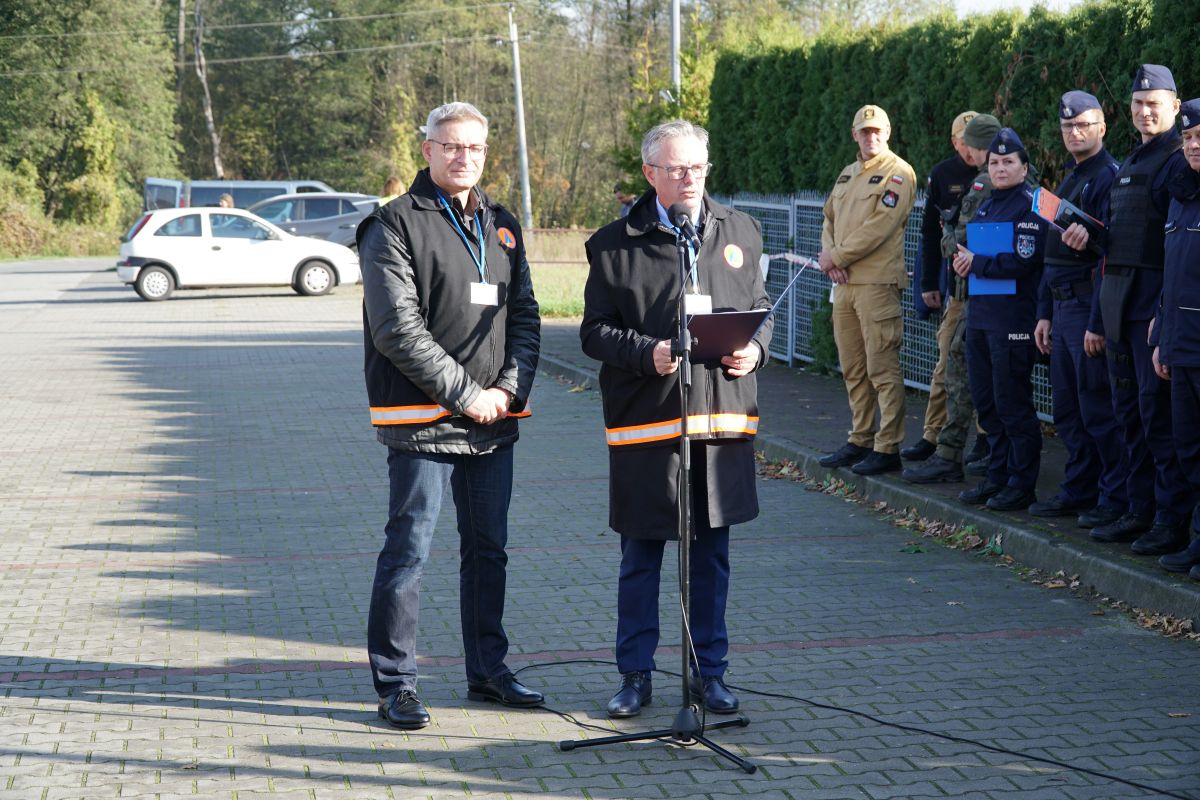 Zdjęcie przedstawia dwóch mężczyzn oraz grupę osób stojących na parkingu podczas ćwiczeń obronnych