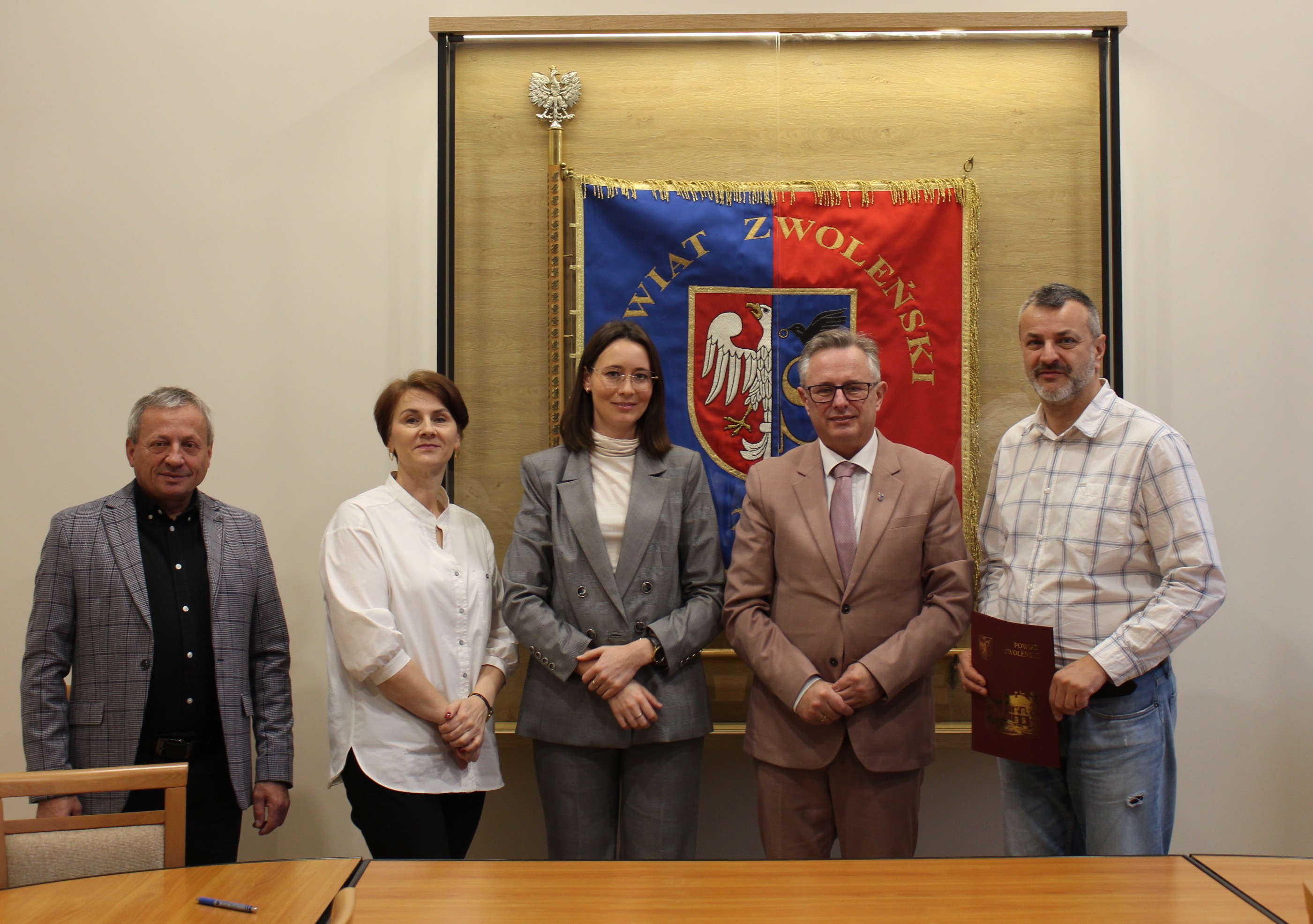 Zdjęcie przedstawia grupę os&oacute;b stojących na tle sztandaru powiatu zwoleńskiego w sali konferencyjnej Starostwa Powiatowego w Zwoleniu
