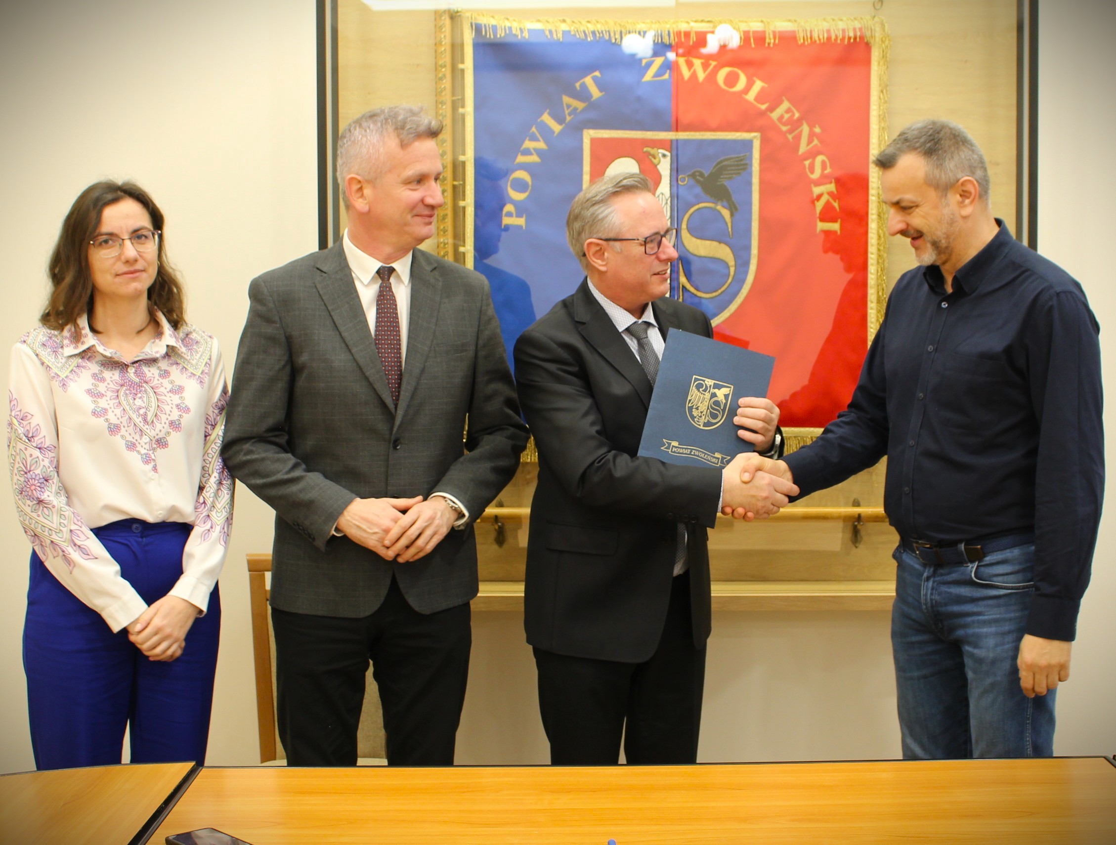 Zdjęcie przedstawia kobietę i trzech mężczyzn na tle sztandaru Powiatu Zwoleńskiego w sali narad Starostwa Powiatowego w Zwoleniu.
