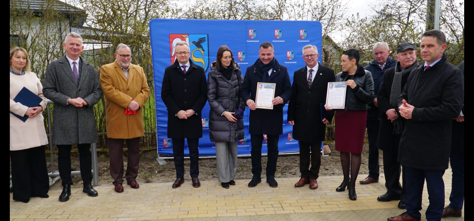 Zdjęcie przedstawia grupę ludzi stojących na chodniku przy drodze. W tle widoczna jest ścianka promocyjna Powiatu Zwoleńskiego.