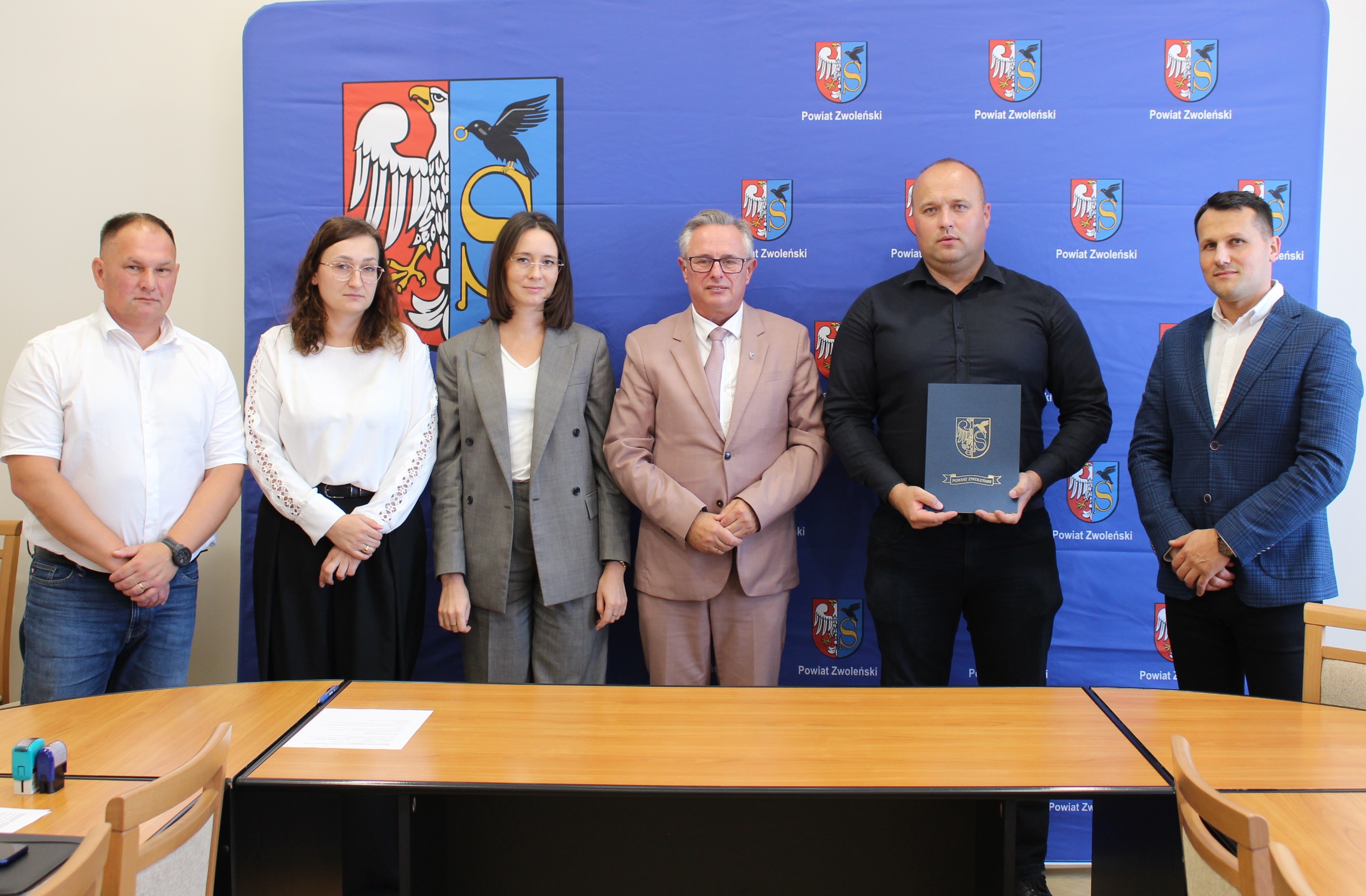 Zdjęcie przedstawia grupę ludzi stojących na ściance promocyjnej Powiatu Zwoleńskiego po podpisaniu umowy.